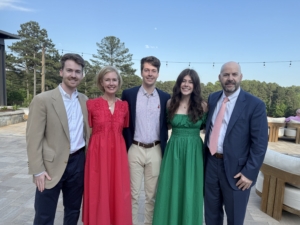 Five people posing for a family photo under a blue sky. They are wearing formal attire and smiling.